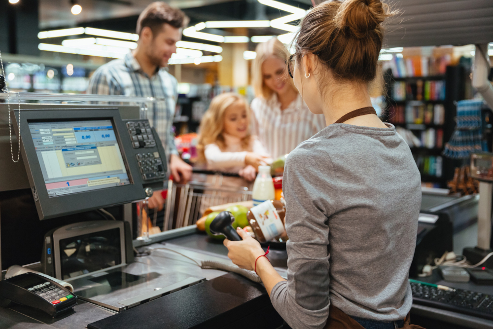Imagem de uma operação de autoatendimento em loja de conveniência, mostrando cliente utilizando terminal de pagamento enquanto uma funcionária acompanha o processo. A cena transmite modernização, tecnologia e transformação no varejo brasileiro.